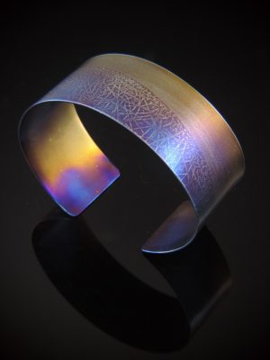 One inch wide titanium cuff with asymmetrical, roll printed line texture torch fired in sunset hues. Approximate size: S/M (fits wrist sizes 6.5 to 7 inches)